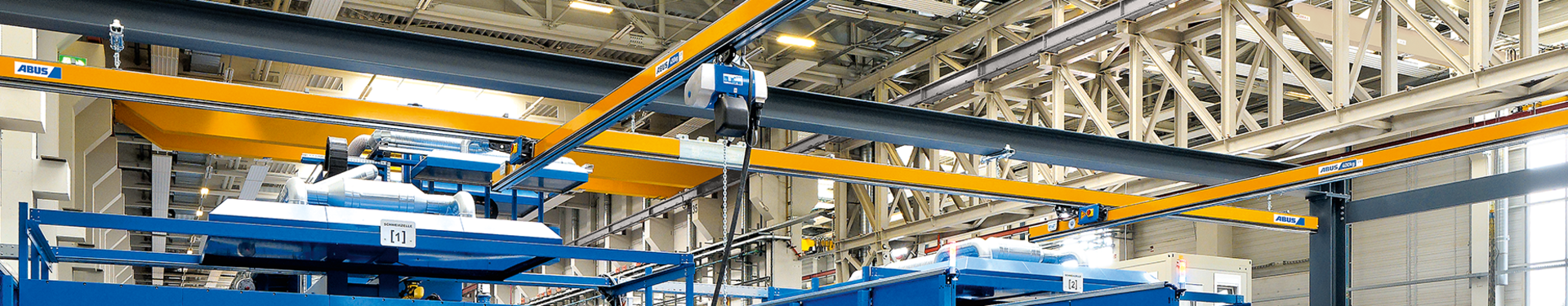 Enkelsteunende hangbaan type EHB In een industriële hal zijn meerdere enkelsteunende hangbanen van het type EHB te zien. Op de voorgrond staat een werknemer die met een hangende schakelaar een kettingtakel bedient. De kettingtakel loopt met een rolwagen in het profiel van de hoofddrager.