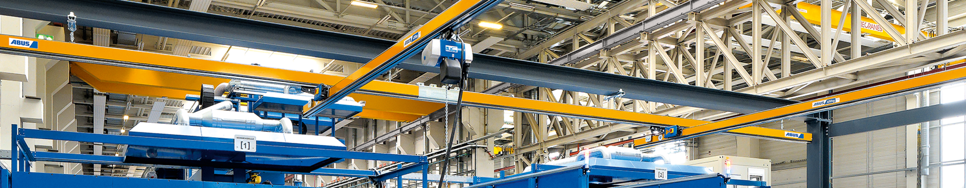 In een industriële hal zijn meerdere enkelsteunende hangbanen van het type EHB te zien. Op de voorgrond staat een werknemer die met een hangende schakelaar een kettingtakel bedient. De kettingtakel loopt met een rolwagen in het profiel van de hoofddrager.