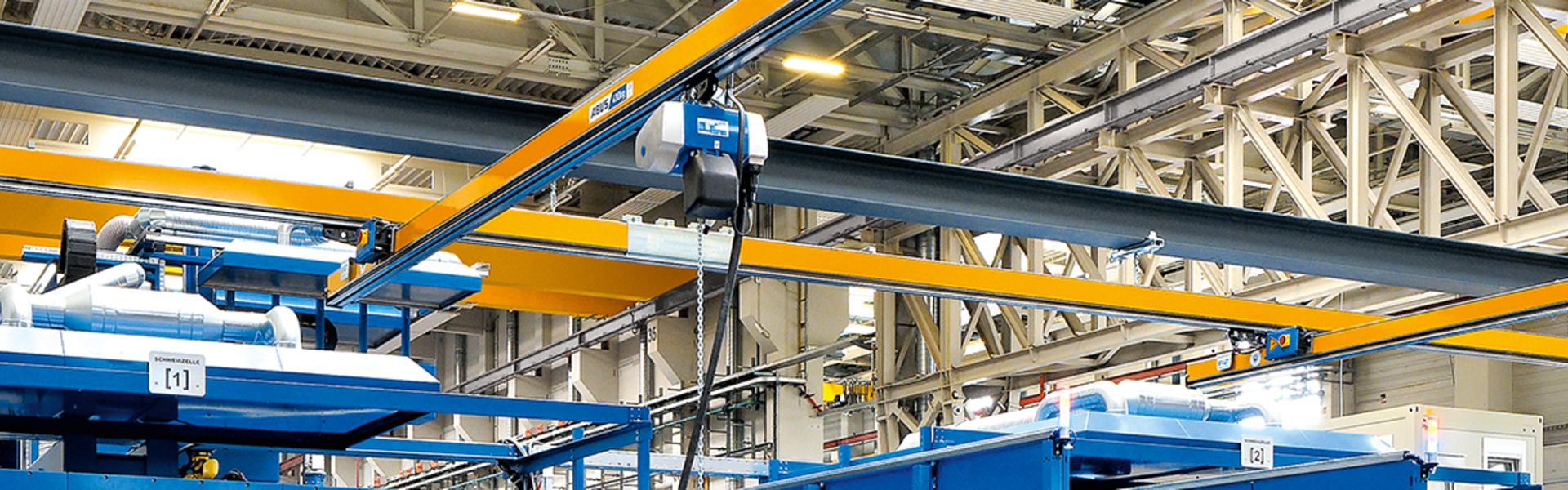 In een industriële hal zijn meerdere enkelsteunende hangbanen van het type EHB te zien. Op de voorgrond staat een werknemer die met een hangende schakelaar een kettingtakel bedient. De kettingtakel loopt met een rolwagen in het profiel van de hoofddrager.