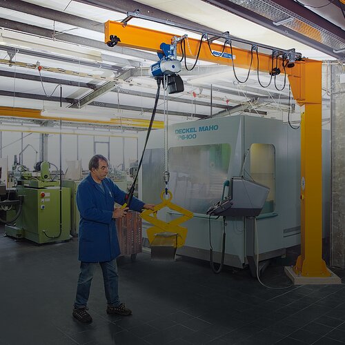 Een medewerker bedient een kolomzwenkkraan en hijst een last met een kettingtakel in een fabriekshal.